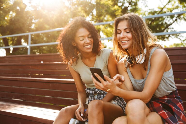 how to manifest a text message - two women sit outside at a bench looking at a phone together and smiling