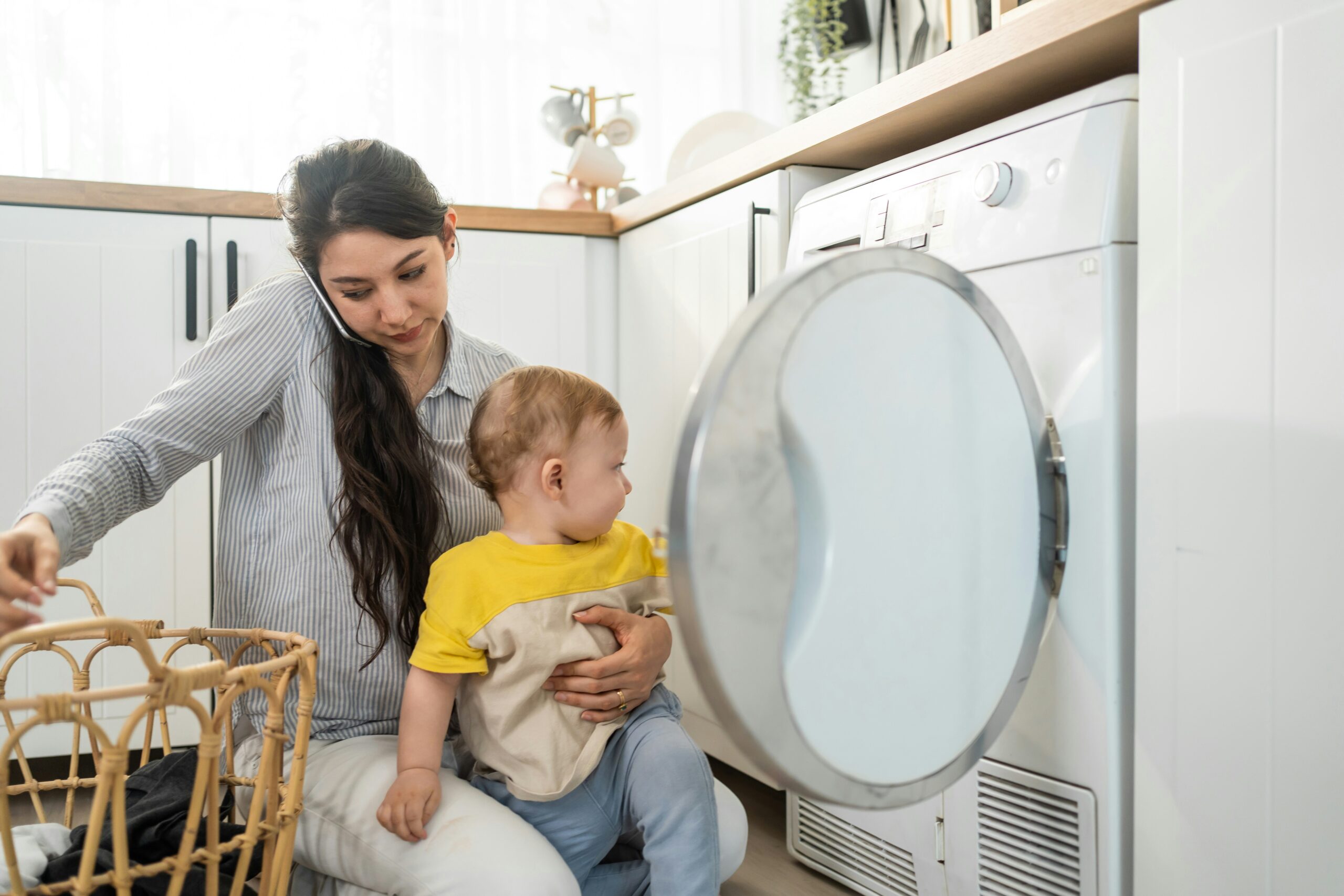 young mother loads laundry into a washer while talking on the phone and holding her toddler